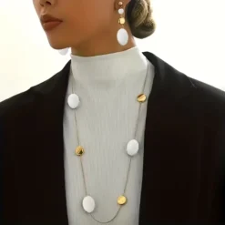 Γυναίκα που φοράει μακρύ χρυσό κολιέ με μαύρα οβάλ στοιχεία και ασορτί κρεμαστά σκουλαρίκια πάνω από μαύρο σακάκι.