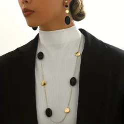 Γυναίκα που φοράει μακρύ χρυσό κολιέ με μαύρα οβάλ στοιχεία και ασορτί κρεμαστά σκουλαρίκια πάνω από μαύρο σακάκι.