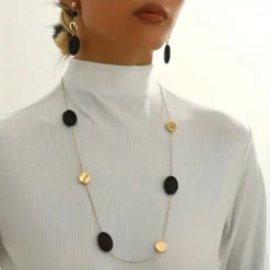 Γυναίκα που φοράει μακρύ χρυσό κολιέ με μαύρα οβάλ στοιχεία και ασορτί κρεμαστά σκουλαρίκια πάνω από μαύρο σακάκι.