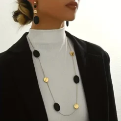 Γυναίκα που φοράει μακρύ χρυσό κολιέ με μαύρα οβάλ στοιχεία και ασορτί κρεμαστά σκουλαρίκια πάνω από μαύρο σακάκι.