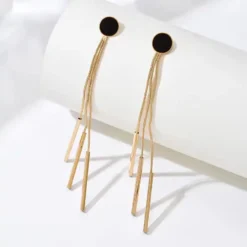 Ζευγάρι μακριά χρυσά γυναικεία σκουλαρίκια με μαύρο κυκλικό στοιχείο και κρεμαστές αλυσίδες σε λευκό φόντο.