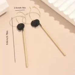 Μακριά γεωμετρικά σκουλαρίκια σε χρυσό χρώμα με μαύρο κυκλικο δίσκο και λεπτή μεταλλική μπάρα, μήκους 9.6 εκατοστών.