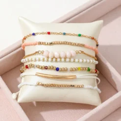 Σετ από επτά γυναικεία βραχιόλια σε boho στυλ με πολύχρωμες χάντρες, χρυσές αλυσίδες και λευκό κορδόνι πάνω σε διακοσμητικό μαξιλαράκι.